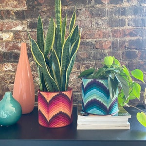 May include: Two potted plants with patterned fabric planters. The plant on the left is a snake plant with green and yellow leaves. The plant on the right is a leafy green plant with a blue and green patterned planter. Both plants are on a black surface with books stacked in front of the plant on the right.