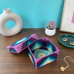 Puede incluir: Una caja cuadrada de tela con un patrón geométrico morado y azul. La caja tiene una tapa.