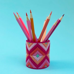 Könnte beinhalten: Ein geometrisch gemusterter Stifthalter in Pink und Orange mit verschiedenen Buntstiften, die oben herausragen.