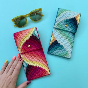 Op de afbeelding: Twee kleurrijke oogstasetuis met geometrische patronen. Een etui is blauw, groen en wit. De andere etui is roze, geel en rood. Beide etuis hebben een gouden knoop.