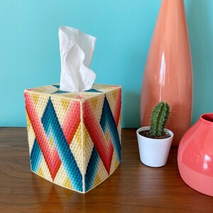 May include: A tissue box cover with a geometric pattern in shades of yellow, pink, blue, and white. The cover is made of needlepoint and has a diamond pattern.