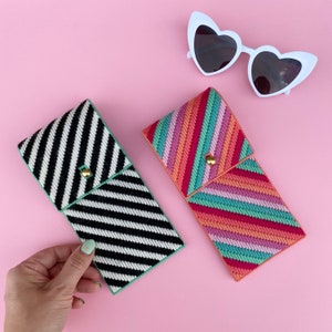 Könnte beinhalten: Zwei gestreifte Brillenetuis aus Stoff. Ein Etui ist schwarz-weiß, das andere ist rosa, grün und weiß. Beide Etuis haben einen goldenen Knopfverschluss.