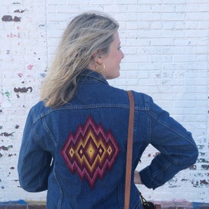 May include: A denim jacket with a colorful geometric design on the back. The design features shades of pink, yellow, and burgundy. The jacket has a classic collar and button-up front.