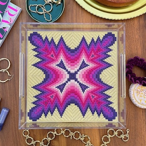 Puede incluir: Bandeja de servicio de acrílico transparente con un diseño de tapiz geométrico vibrante en tonos de morado, rosa y amarillo. La bandeja tiene dos asas. Una cadena dorada es visible en la parte inferior.