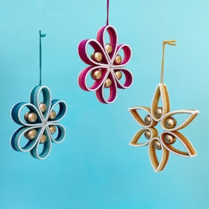 Pode incluir: Três ornamentos feitos à mão em forma de flores e uma estrela. Os ornamentos são feitos de fio e têm contas douradas no centro de cada forma. Os ornamentos estão pendurados em um fundo azul.