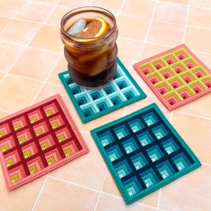 May include: A glass of iced tea with an orange slice and ice cubes sits on a square, colorful, needlepoint coaster. Three additional coasters in shades of pink, yellow, and teal are also visible. The coasters have a geometric design.