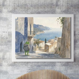 Könnte beinhalten: Ein gerahmtes Aquarellbild, das einen grünen Frosch mit grauem Kapuzenpullover auf einem Steinweg zeigt. Die Szene blickt auf eine Küstenstadt mit weißen Gebäuden, einem blauen Meer und fernen Bergen unter einem klaren Himmel.