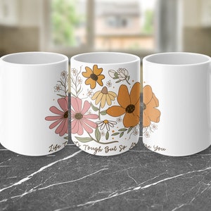 Könnte beinhalten: Weiße Keramiktasse mit einem Blumendesign in Rosa, Orange und Gelb. Auf der Tasse steht der Text "Life is Tough But So Are You". Die Tasse hat einen Standardgriff.