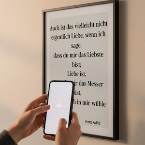 Könnte beinhalten: Ein gerahmter Druck mit schwarzen Rändern zeigt ein Zitat von Franz Kafka in schwarzem Text auf hellem Hintergrund. Ein Smartphone mit weißem Bildschirm wird vor dem Druck gehalten.