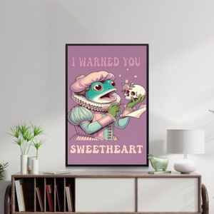 Könnte beinhalten: Ein lilafarbener Poster mit schwarzem Rahmen zeigt einen Comic-Frosch, der ein rosa-blaues Kleid trägt und einen Schädel hält. Der Text auf dem Poster lautet "Ich habe dich gewarnt, mein Schatz."