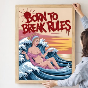 Könnte beinhalten: Ein gerahmter Kunstdruck mit dem Text "BORN TO BREAK RULES" in Rot über einer Sonnenuntergangsszene. Das Kunstwerk zeigt eine Frau in einem rosa Kleid, die in einer großen Meereswelle sitzt. Der Druck ist in einem Holzrahmen.