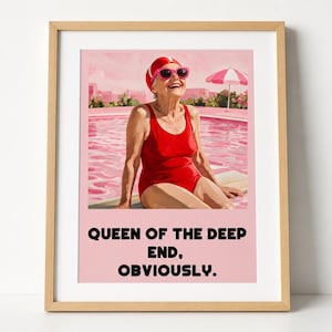 Könnte beinhalten: Ein gerahmter Kunstdruck mit einer älteren Person in rotem Badeanzug, roter Badekappe und rosa Sonnenbrille am Rand eines rosa Swimmingpools mit gestreiftem Sonnenschirm. Der Text lautet: QUEEN OF THE DEEP END, OBVIOUSLY. Der Rahmen ist aus hellem Holz.