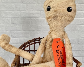 May include: A beige mummy doll wrapped in textured fabric strips with black button eyes, sitting on a small rustic twig chair. An orange rectangular tag hangs from the doll's neck, featuring black text that reads Raggedy Mummy with stars.