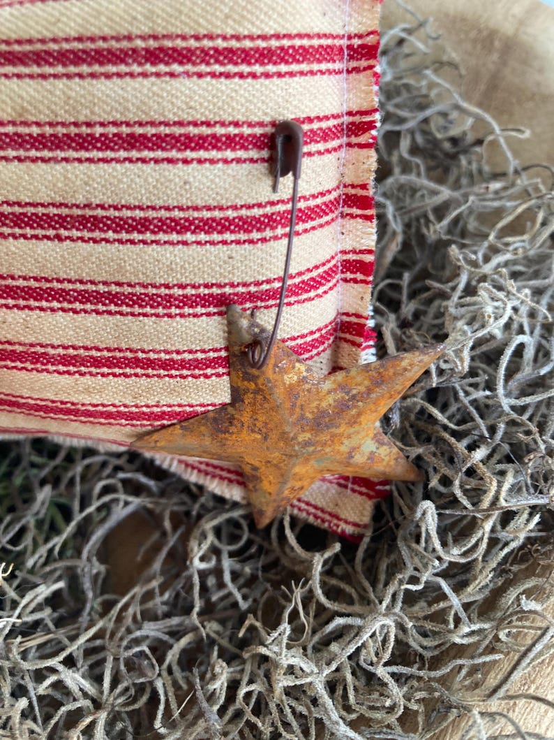 Puede incluir: Primer plano de un adorno met&aacute;lico en forma de estrella oxidada, con acabado naranja. La estrella est&aacute; sujeta a una tela crema con rayas rojas con un imperdible. El fondo presenta material vegetal seco, de tono gris, creando un ambiente vintage.