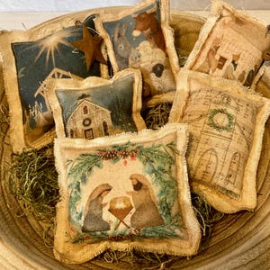May include: A collection of small, burlap-edged pillows with vintage-style Christmas scenes, including a nativity scene, church, and star. The pillows are arranged in a wooden bowl filled with green moss, creating a rustic holiday display.
