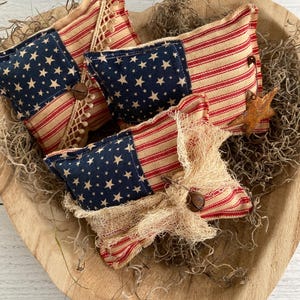 Puede incluir: Una colecci&oacute;n de cojines decorativos con tem&aacute;tica patri&oacute;tica. Los cojines presentan una combinaci&oacute;n de tela azul con estrellas blancas y tela a rayas rojas y beige. Algunos cojines est&aacute;n adornados con botones de madera y detalles de arpillera, dispuestos en un cuenco de madera.