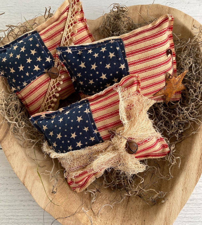 Puede incluir: Una colecci&oacute;n de cojines decorativos con tem&aacute;tica patri&oacute;tica. Los cojines presentan una combinaci&oacute;n de tela azul con estrellas blancas y tela a rayas rojas y beige. Algunos cojines est&aacute;n adornados con botones de madera y detalles de arpillera, dispuestos en un cuenco de madera.