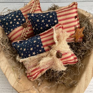 Puede incluir: Un cuenco de madera contiene varias almohadas decorativas de tem&aacute;tica patri&oacute;tica. Las almohadas presentan una combinaci&oacute;n de rayas rojas y blancas y tela azul con estrellas blancas, que recuerdan a la bandera americana. Algunas almohadas est&aacute;n adornadas con encaje, campanas y detalles r&uacute;sticos.