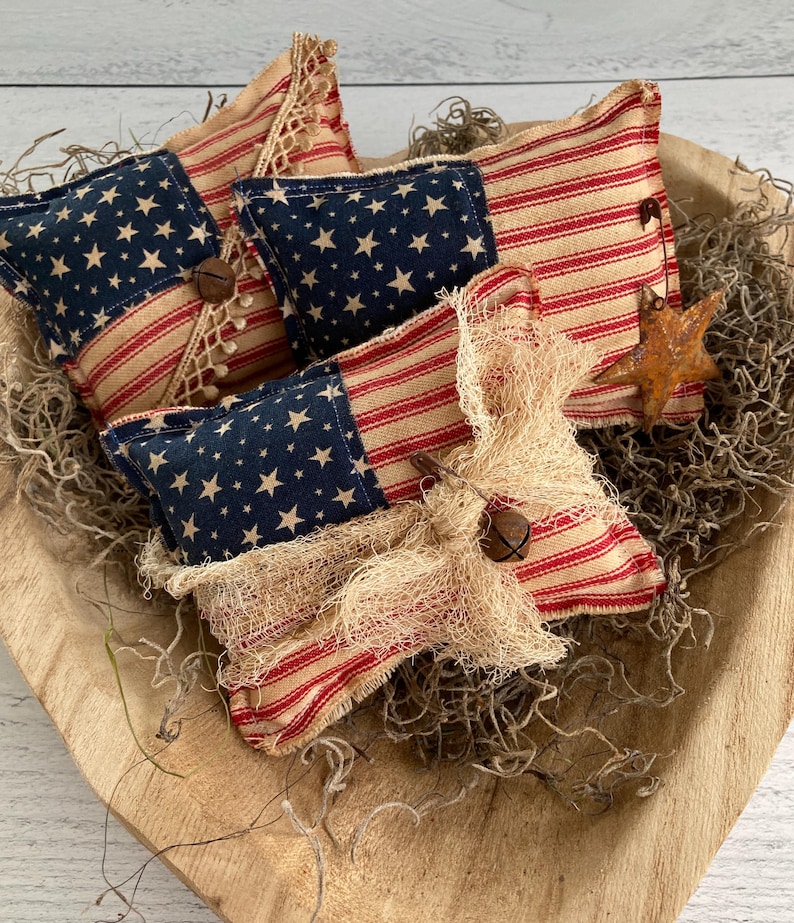 Puede incluir: Un cuenco de madera contiene varias almohadas decorativas de tem&aacute;tica patri&oacute;tica. Las almohadas presentan una combinaci&oacute;n de rayas rojas y blancas y tela azul con estrellas blancas, que recuerdan a la bandera americana. Algunas almohadas est&aacute;n adornadas con encaje, campanas y detalles r&uacute;sticos.