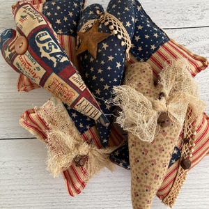 Puede incluir: Una colecci&oacute;n de adornos de tela en forma de coraz&oacute;n con tem&aacute;tica patri&oacute;tica. Los adornos presentan patrones de estrellas y rayas, junto con texto de estilo vintage. Est&aacute;n decorados con elementos r&uacute;sticos como encaje, arpillera y botones de madera. La paleta de colores incluye azul marino, rojo y beige.