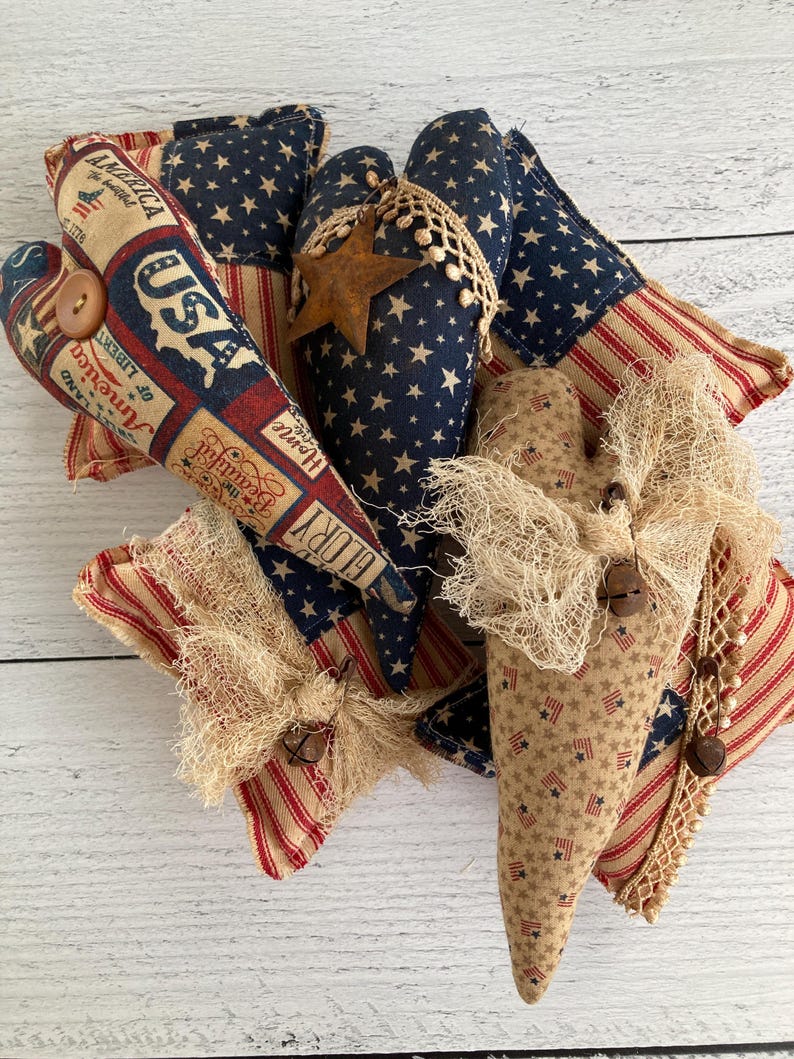 Puede incluir: Una colecci&oacute;n de adornos de tela en forma de coraz&oacute;n con tem&aacute;tica patri&oacute;tica. Los adornos presentan patrones de estrellas y rayas, junto con texto de estilo vintage. Est&aacute;n decorados con elementos r&uacute;sticos como encaje, arpillera y botones de madera. La paleta de colores incluye azul marino, rojo y beige.