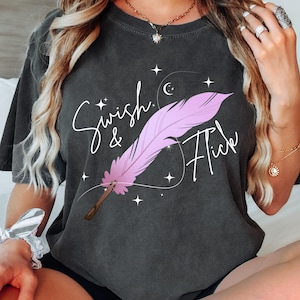 May include: Dark gray t-shirt with the words "Swish & Flick" and a pink feather design. The design includes stars and a crescent moon. The shirt is short-sleeved and has a relaxed fit.