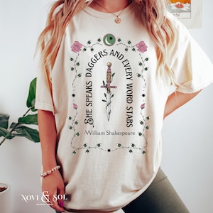 Puede incluir: Camiseta color crema con un dise&ntilde;o decorativo que presenta una daga, rosas y la cita "She speaks daggers and every word stabs" de William Shakespeare. El dise&ntilde;o incluye una luna creciente y estrellas.
