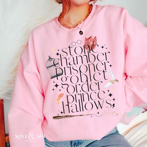 Puede incluir: Una sudadera rosa claro con un diseño gráfico que presenta las palabras "Stone Chamber Prisoner Goblet Order Prince Hallows" en una fuente estilizada. El diseño incluye ilustraciones de una jaula, gafas y una varita.