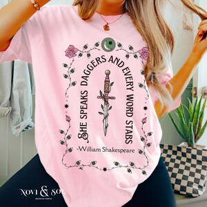 Puede incluir: Camiseta rosa claro con un dise&ntilde;o gr&aacute;fico. El dise&ntilde;o incluye una daga, rosas y la cita "She speaks daggers and every word stabs" de William Shakespeare. El nombre de la marca "Novi & So" est&aacute; en la parte inferior. La camiseta tiene cuello redondo.