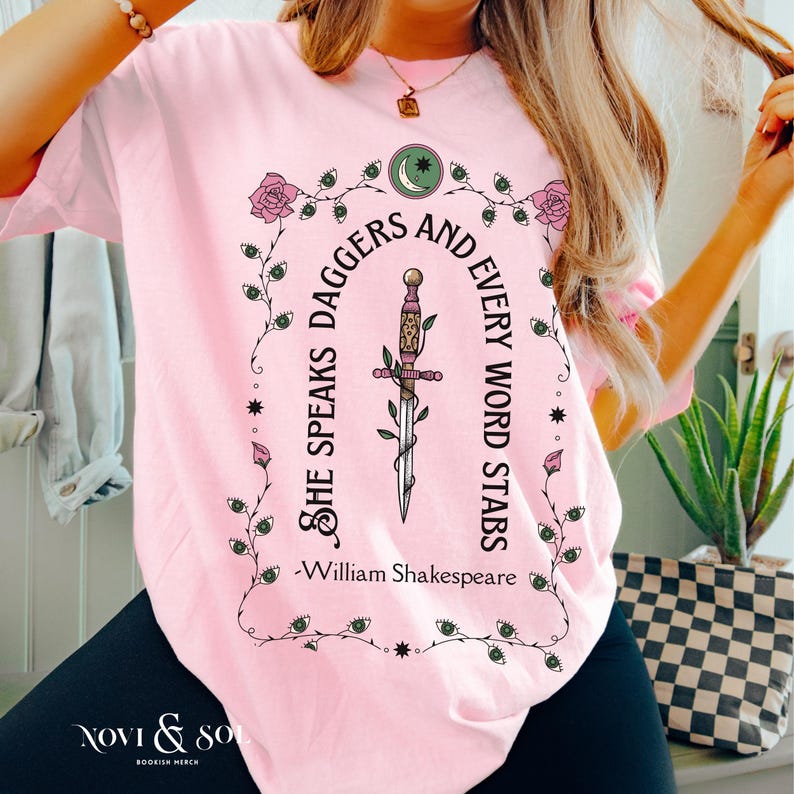 Puede incluir: Camiseta rosa claro con un dise&ntilde;o gr&aacute;fico. El dise&ntilde;o incluye una daga, rosas y la cita "She speaks daggers and every word stabs" de William Shakespeare. El nombre de la marca "Novi & So" est&aacute; en la parte inferior. La camiseta tiene cuello redondo.