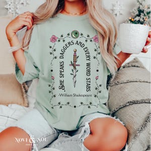 Puede incluir: Camiseta verde claro con la cita "She Speaks Daggers and Every Word Stars" y una ilustraci&oacute;n de daga. El dise&ntilde;o incluye rosas rosas y una luna creciente. El texto "William Shakespeare" est&aacute; debajo.