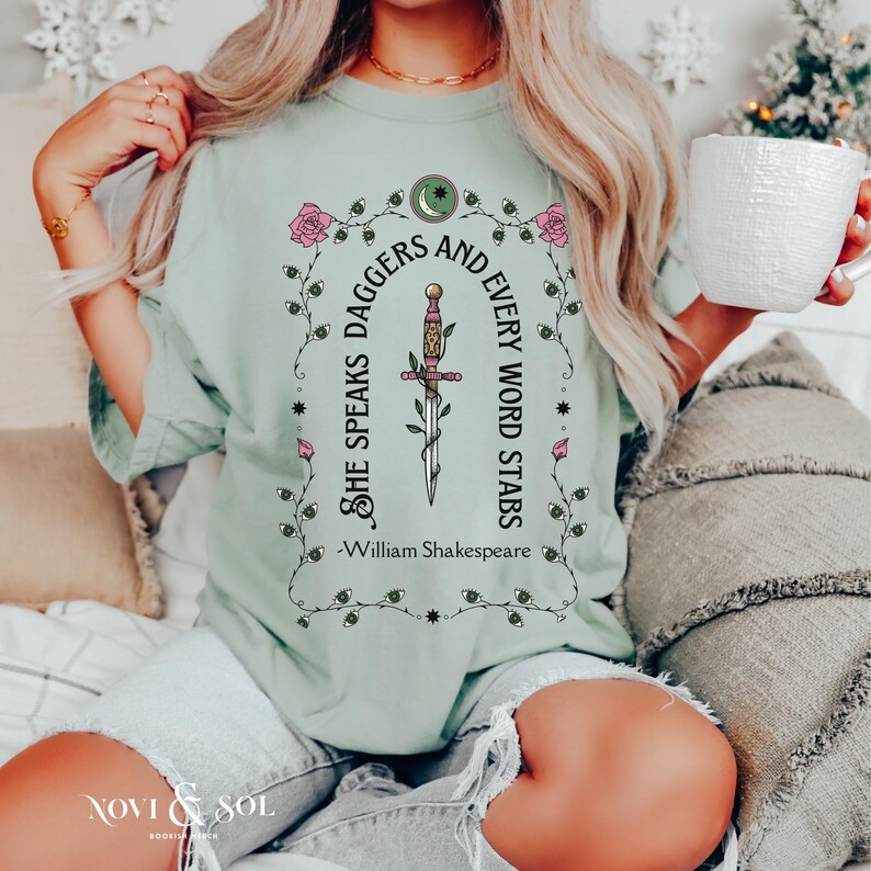 Puede incluir: Camiseta verde claro con la cita "She Speaks Daggers and Every Word Stars" y una ilustraci&oacute;n de daga. El dise&ntilde;o incluye rosas rosas y una luna creciente. El texto "William Shakespeare" est&aacute; debajo.