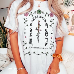 Puede incluir: Camiseta blanca con un gr&aacute;fico que presenta una daga y la cita "She Speaks Daggers and Every Word Stabs" de William Shakespeare. El dise&ntilde;o incluye elementos florales y celestiales en verde y rosa.