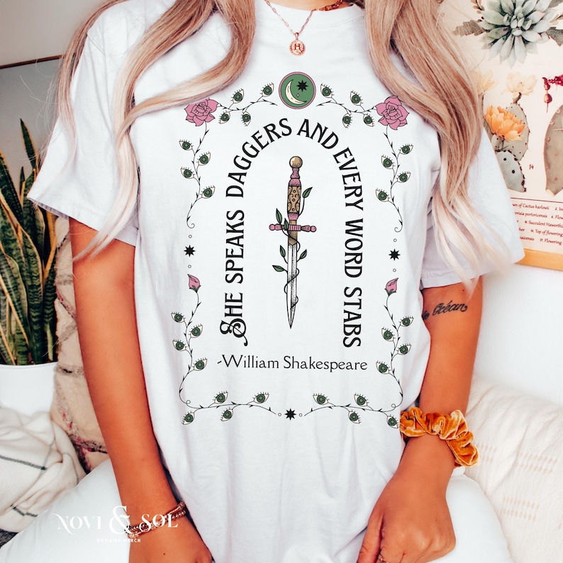 Puede incluir: Camiseta blanca con un gr&aacute;fico que presenta una daga y la cita "She Speaks Daggers and Every Word Stabs" de William Shakespeare. El dise&ntilde;o incluye elementos florales y celestiales en verde y rosa.