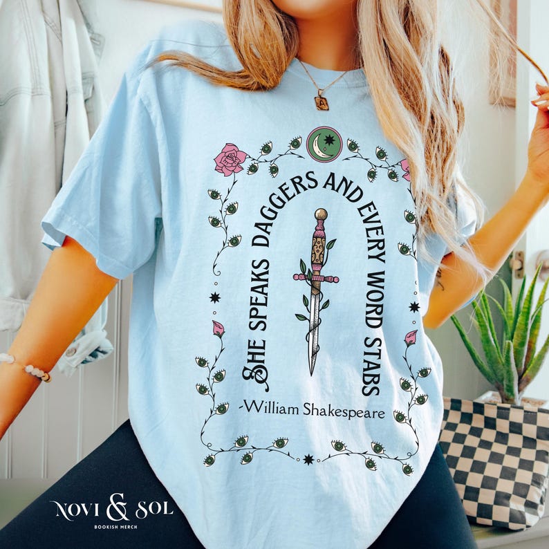 Puede incluir: Camiseta azul claro con un gr&aacute;fico que presenta una daga, rosas y la cita "She speaks daggers and every word stabs" de William Shakespeare. La camiseta tambi&eacute;n tiene la marca "Novi & Sol" en la parte inferior.