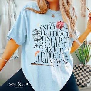 May include: Light blue t-shirt with a graphic featuring the words "stone chamber prisoner goblet order prince hallows" in a stylized font. The design includes a birdcage, glasses, and a wand. The shirt has short sleeves.
