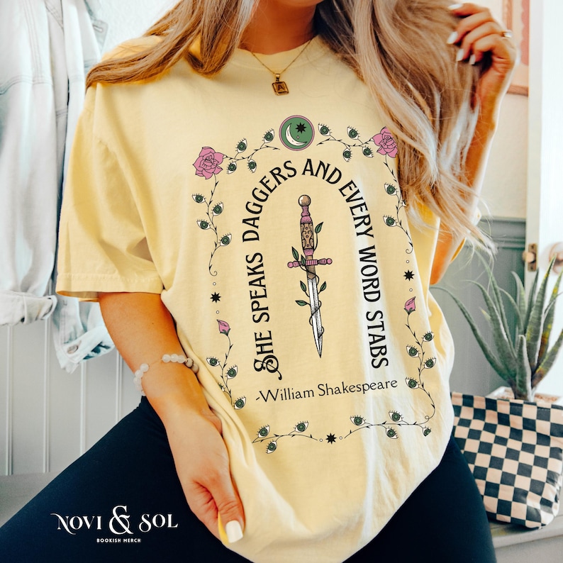 Puede incluir: Camiseta amarilla clara con un dise&ntilde;o literario. El gr&aacute;fico incluye una daga y la cita "She Speaks Daggers and Every Word Stabs" de William Shakespeare, con elementos florales. Tambi&eacute;n se ve la marca "Novi & Sol".