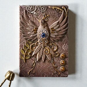 Pinky Celestial Bird & Phoenix cover Journal, 3D embossed fantasy notebook with silver cover, wings-spread phoenix design, blue gemstone and gold accents, intricate floral and swirling patterns, three gold gems at the bottom. Lined pages, A5 size. Fantasy journal, enchanted diary, whimsical notebook, magical writing notebook, decorative collectible, gift for writers, dreamers, and fantasy lovers, mystical aesthetic. Perfect gift for readers and book lovers.