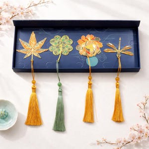 Peut inclure: Un ensemble de quatre marque-pages décoratifs en métal dans une boîte bleu marine. Les marque-pages présentent un motif de feuille d'érable, de trèfle à quatre feuilles, de fleur et de libellule, chacun avec un pompon. Les pompons sont dorés et verts.