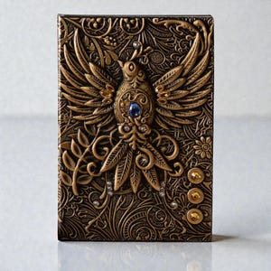 Gold Celestial Bird & Phoenix cover Journal, 3D embossed fantasy notebook with silver cover, wings-spread phoenix design, blue gemstone and gold accents, intricate floral and swirling patterns, three gold gems at the bottom. Lined pages, A5 size. Fantasy journal, enchanted diary, whimsical notebook, magical writing notebook, decorative collectible, gift for writers, dreamers, and fantasy lovers, mystical aesthetic. Perfect gift for readers and book lovers.