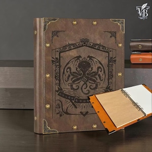 May include: Brown leather journal with an engraved octopus design, brass corner details, and a ring binder. The journal's cover features an intricate design with wings and geometric shapes. A partially open journal with blank pages is visible.