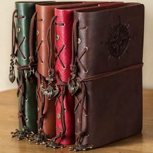 Op de afbeelding: Een verzameling lederen journals in verschillende kleuren: groen, bruin, rood en donkerbruin. Elk journal heeft een decoratieve bedel en een kompasroosontwerp op de voorkant. De journals zijn vastgebonden met een leren koord.