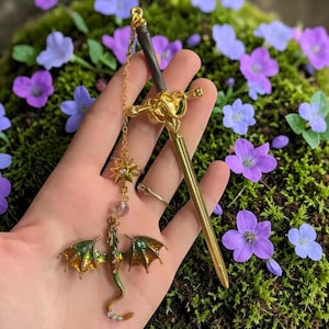 Peut inclure: Un accessoire pour cheveux en forme d'épée dorée avec un charm en forme de dragon décoratif. Le dragon a des ailes vertes et dorées et un long corps sinueux. Une étoile et des perles roses pendent de l'épée. Le bâton à cheveux mesure environ 18 cm de long.
