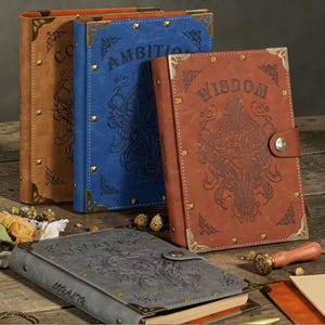Op de afbeelding: Een verzameling lederen journals in bruin, blauw, rood en grijs. Elk journal heeft reliëfontwerpen, hoekbeschermers en de woorden "Ambition", "Wisdom", "Loyalty" en "Courage" op de covers. De journals hebben een vintage esthetiek.