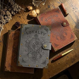 Op de afbeelding: Drie lederen journals met gouden accenten en decoratieve hoekstukken. De journals zijn in bruin- en grijstinten, met de woorden "Loyalty", "Wisdom" en "Co" in reliëf op de omslagen. Een gouden pen en een zegel zijn ook zichtbaar.