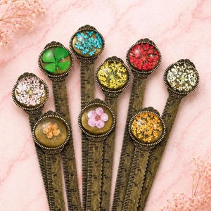 Peut inclure: Une collection de marque-pages en métal bronze antique, chacun avec une fleur ou une plante séchée différente enfermée dans un dôme de résine transparente. Les marque-pages ont un design de règle et sont disposés sur une surface rose.