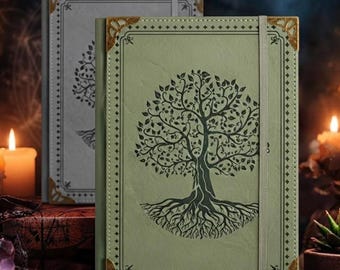 May include: A sage green journal featuring a dark tree of life illustration on the cover, accented with gold-toned metal corner protectors and a vertical elastic closure band. A second gray journal with a matching design is positioned behind it.