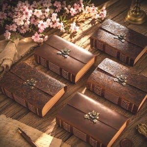 Puede incluir: Cinco diarios de cuero marrón con cierres ornamentados y patrones florales en relieve están dispuestos sobre una superficie de madera. Los diarios tienen páginas envejecidas y texturizadas, acentuados por un bolígrafo, flores y un sello de cera, creando una estética vintage.