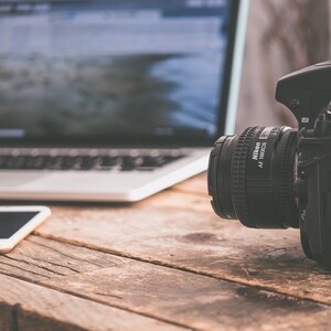 May include: A black DSLR camera with a large lens sits on a wooden table next to a laptop and a smartphone.