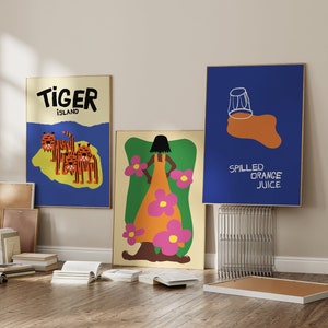 Può includere: Tre illustrazioni colorate incorniciate e appese a un muro. La prima illustrazione è uno sfondo blu con un'isola gialla e il testo "Tiger Island". La seconda illustrazione è uno sfondo giallo con una donna in un vestito rosa e arancione. La terza illustrazione è uno sfondo blu con un bicchiere di succo d'arancia e il testo "Spilled Orange Juice".